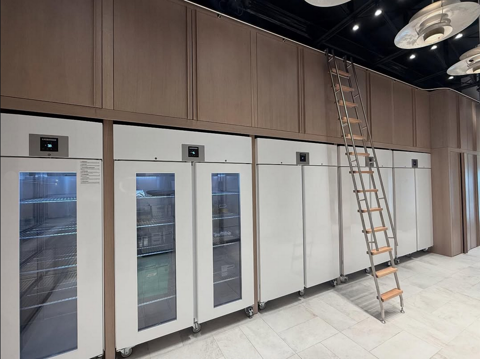 Professional kitchen featuring a row of glass-door refrigerators, custom wood upper cabinetry, and a rolling stainless steel library ladder.