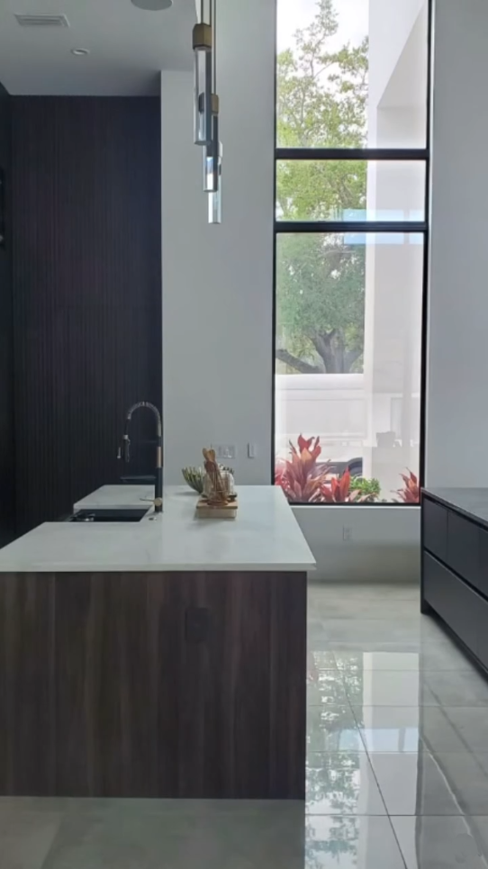 Modern kitchen island with dark wood paneling and white marble top, featuring a large window with garden views and designer pendant lighting.