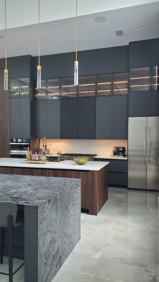Modern dark-themed kitchen with matte black cabinets, integrated lighting, a marble waterfall island, and a wood-slat secondary island.