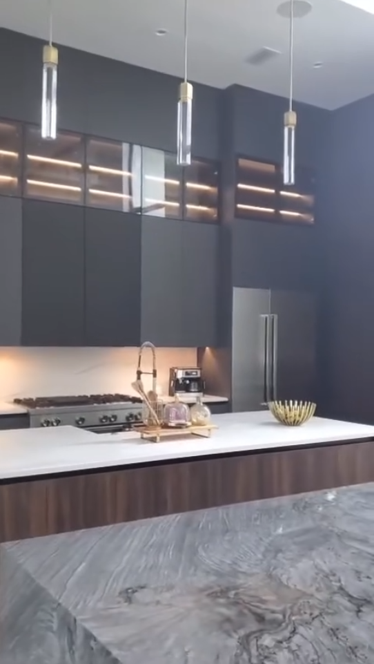 Modern kitchen design featuring grey marble waterfall island, charcoal custom cabinets, backlit glass display cases, and professional stainless steel appliances.