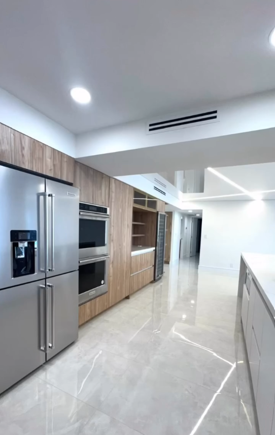 Modern kitchen with natural wood cabinets, integrated stainless steel appliances, and white high-gloss flooring with linear LED accents.