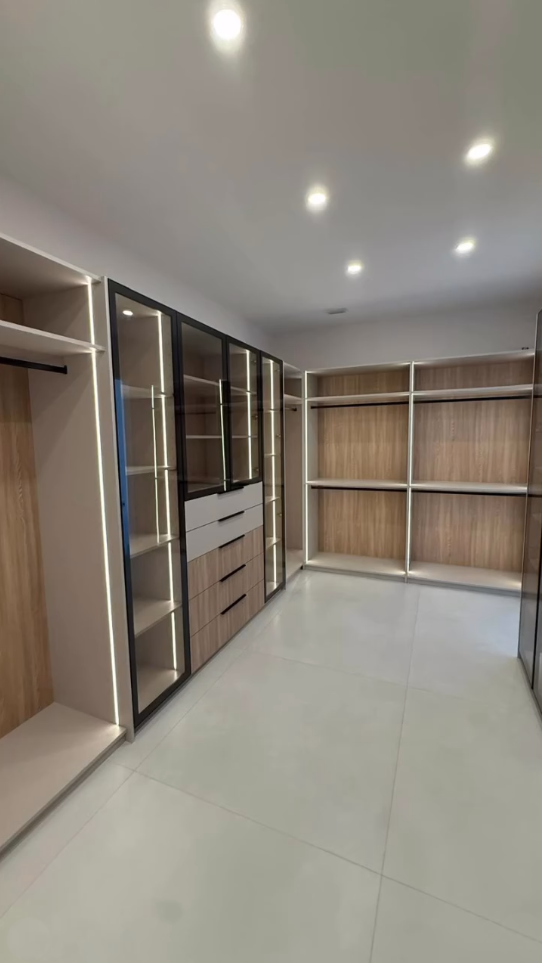 Modern luxury walk-in closet featuring integrated vertical LED lighting, light wood cabinetry, and black-framed glass display cases.
