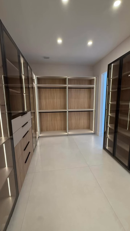 Modern luxury walk-in closet featuring integrated vertical LED lighting, light wood cabinetry, and black-framed glass display cases.