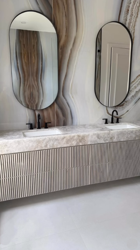 Luxury bathroom design featuring book-matched stone wall, and oval mirrors with black hardware.