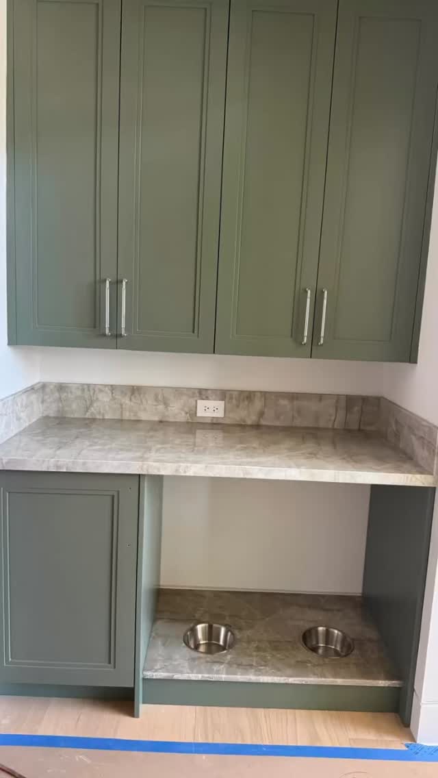 Custom sage green cabinetry featuring a built-in pet feeding station. The unit includes a stone countertop and a recessed floor-level nook with two stainless steel bowls integrated into a matching stone base.